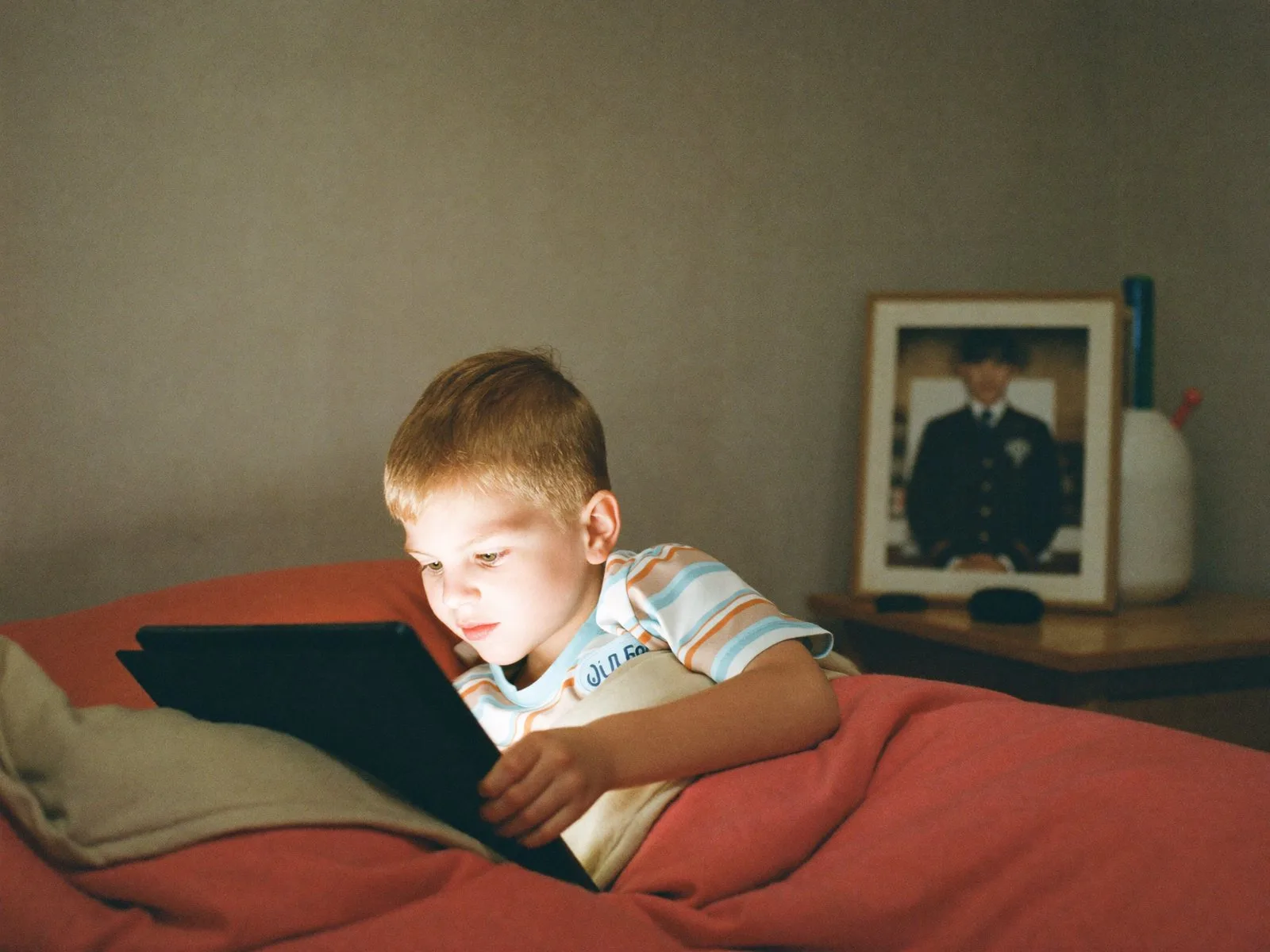 A child in pyjamas holding a tablet in a dark bedroom, a framed photo of a parent in uniform beside the bed