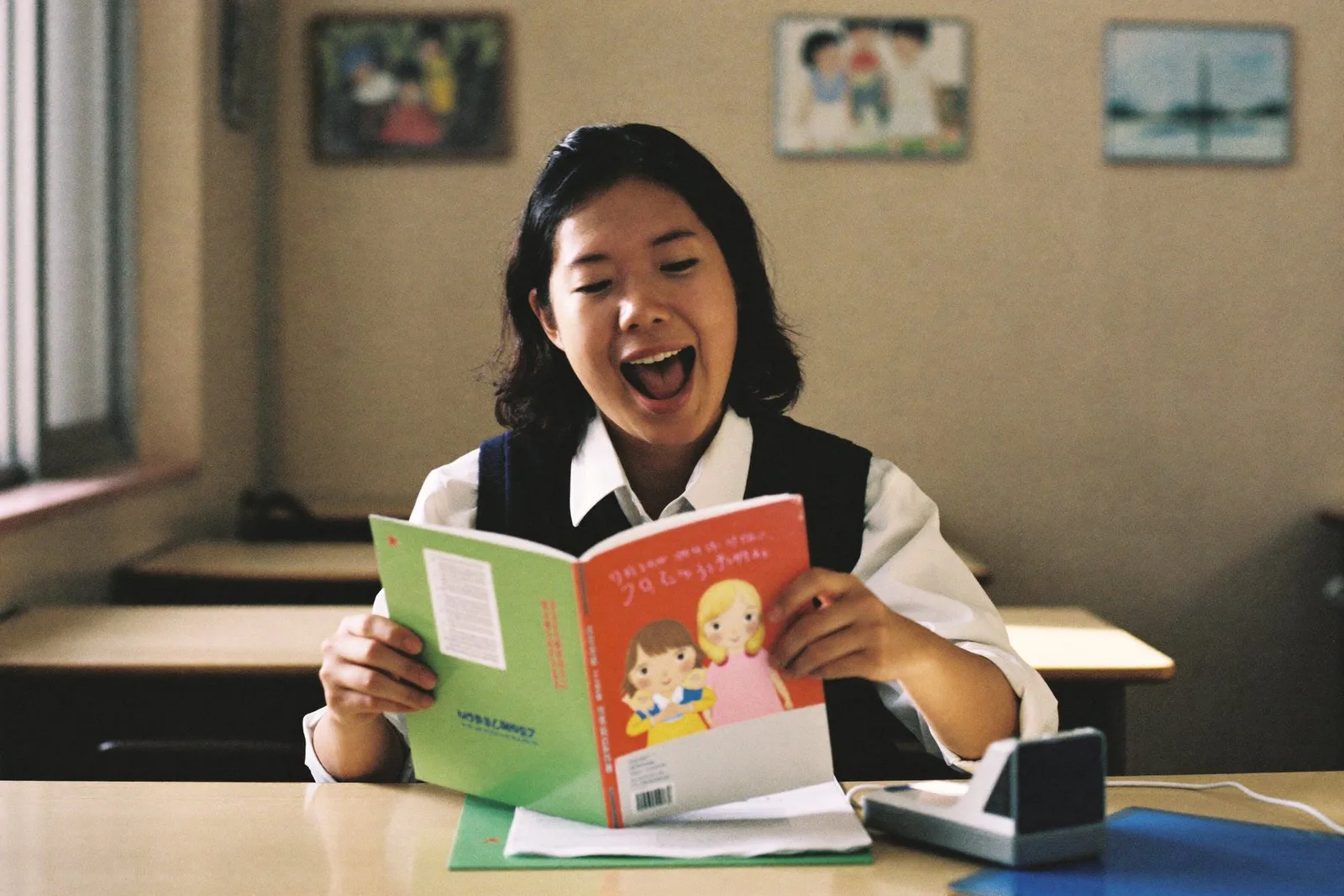 A teacher at her desk in an empty classroom, reading a picture book aloud to her phone