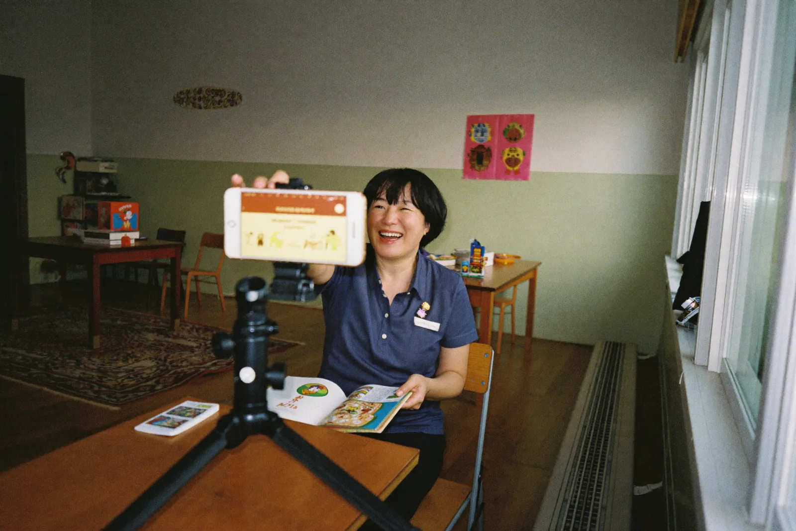 A teacher at her desk in an empty classroom, reading a picture book aloud to her phone