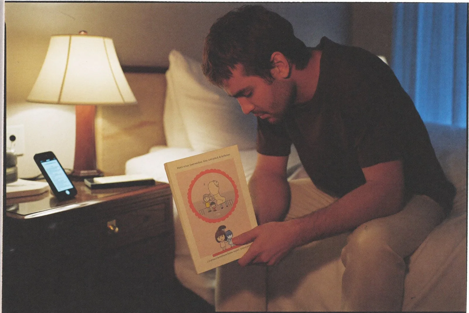 A father reading a picture book aloud toward his phone in a hotel room at night